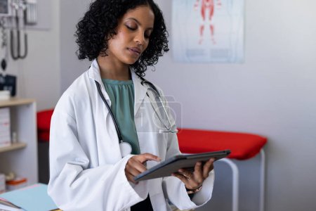 Foto de Médico Biracial femenino usando estetoscopio, usando tableta en el consultorio del médico. Hospital, medicina, salud y comunicación. - Imagen libre de derechos