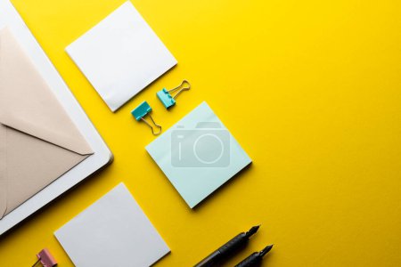 Flat lay of black pens, envelope, paper clips and memo notes with copy space on yellow background. School materials, writing, colouring, drawing, learning, school and education concept.