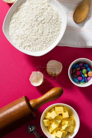 Photo for Directly above shot of flour, butter and colorful candies in bowls, rolling pin, spoon and napkin. pink background, copy space, eggshell, spoon, food, cookie, ingredients and preparation concept. - Royalty Free Image