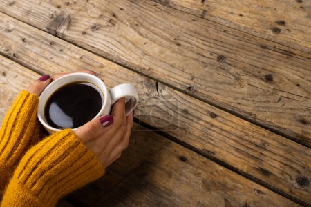 Photo for High angle cropped hands of caucasian woman wearing yellow sweater holding coffee cup on table. Wood, unaltered, drink, caffeine, winter concept. - Royalty Free Image