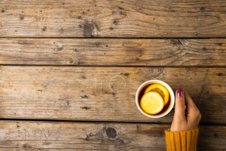 Téléchargez les photos : Main coupée de femme caucasienne portant un pull jaune tenant une tasse de thé avec une tranche de citron sur la table. Bois, vue aérienne, intact, agrumes, boisson et concept d'hiver. - en image libre de droit