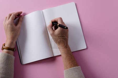 Photo for Hands of caucasian woman holding pen and writing in notebook with copy space on pink background. Literature, writing, leisure time and books. - Royalty Free Image