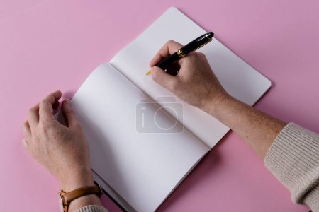 Foto de Manos de mujer caucásica sosteniendo pluma y escribiendo en cuaderno con espacio de copia sobre fondo rosa. Literatura, escritura, tiempo libre y libros. - Imagen libre de derechos