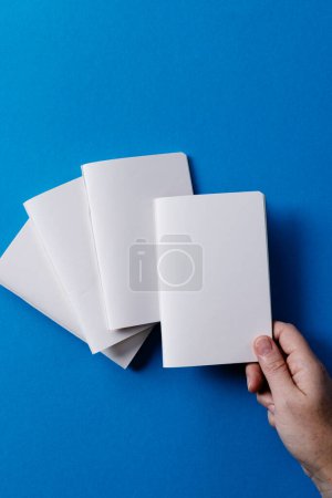 Photo for Hand of caucasian woman holding a notebook over other notebooks with copy space on blue background. Literature, reading, leisure time and books. - Royalty Free Image