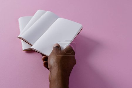 Foto de Mano de hombre biracial sosteniendo cuaderno sobre cuadernos con espacio de copia sobre fondo rosa. Literatura, lectura, tiempo libre y libros. - Imagen libre de derechos