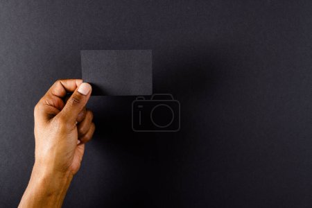 Photo for Hand of biracial man holding black business card with copy space on black background. Business, business card, stationery and writing space concept. - Royalty Free Image
