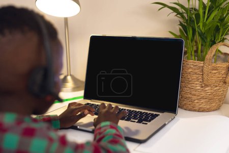 Téléchargez les photos : African american boy in headphones typing on laptop keyboard while studying at home, copy space. Unaltered, childhood, technology, education, student, e-learning, screen, online. - en image libre de droit