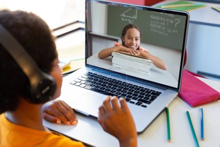 Téléchargez les photos : Biracial boy wearing headphones and discussing homework with girl over video call on laptop. Unaltered, childhood, technology, education, screen, online, student, e-learning and home concept. - en image libre de droit