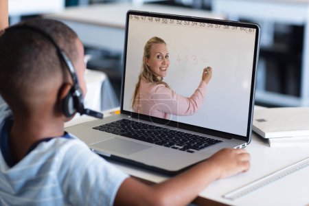Téléchargez les photos : Un garçon afro-américain dans un casque écoutant une enseignante enseigner par appel vidéo sur un ordinateur portable. Inchangé, enfance, technologie, éducation, étudiant, e-learning, en ligne, écran et concept de la maison. - en image libre de droit