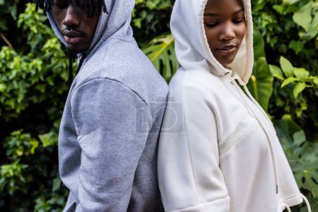 Téléchargez les photos : Couple afro-américain portant des sweats à capuche blancs et gris debout dos à dos dans le jardin. Mode, vêtements décontractés et vêtements de loisir, inchangés. - en image libre de droit