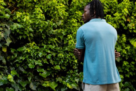 Foto de Vista trasera del hombre afroamericano con rastas que usan polo azul pálido en el jardín, espacio para copiar. Moda, ropa casual y ropa de ocio, inalterada. - Imagen libre de derechos