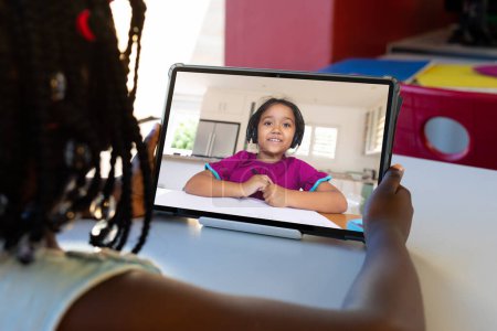 Téléchargez les photos : Vue arrière d'une fille afro-américaine ayant un appel vidéo sur tablette avec une fille biraciale à l'école. Concept d'enseignement en ligne à distance - en image libre de droit