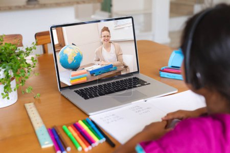 Téléchargez les photos : Vue arrière d'une fille biraciale ayant un appel vidéo avec une enseignante caucasienne sur un ordinateur portable à la maison. Concept d'enseignement en ligne à distance - en image libre de droit