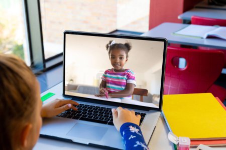 Téléchargez les photos : Fille caucasienne utilisant un ordinateur portable tout en ayant un appel vidéo avec une fille afro-américaine à l'école. Concept d'enseignement en ligne à distance - en image libre de droit