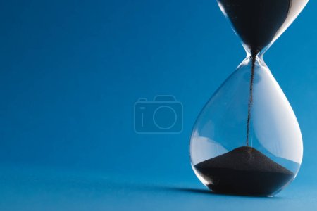 Photo for Close up of hourglass with black sand and copy space on blue background. Time, timekeeping, shape and colour concept. - Royalty Free Image