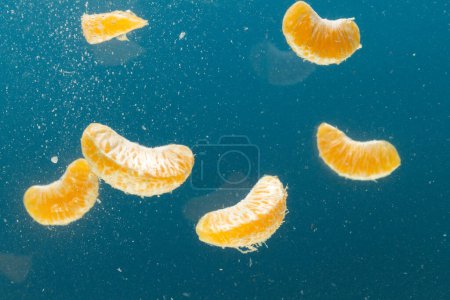 Téléchargez les photos : Gros plan de segments de mandarine tombant dans l'eau avec espace de copie sur fond bleu. Fruits, nourriture végétalienne et concept de couleur. - en image libre de droit
