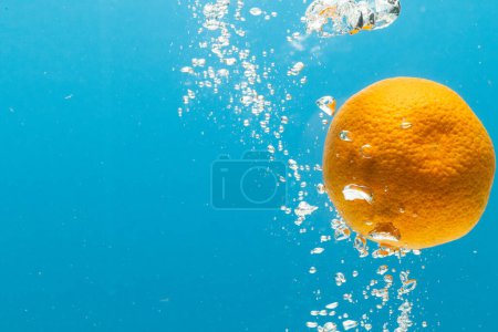 Téléchargez les photos : Gros plan de mandarine tombant dans l'eau avec espace de copie sur fond bleu. Fruits, nourriture végétalienne et concept de couleur. - en image libre de droit