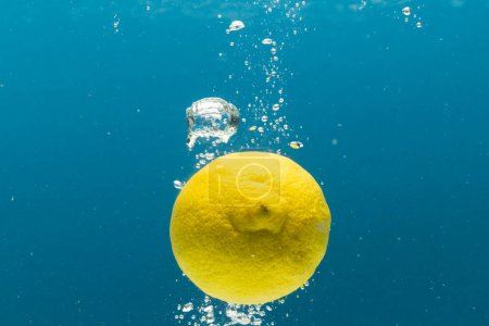 Téléchargez les photos : Gros plan de citron tombant dans l'eau avec un espace de copie sur fond bleu. Fruits, nourriture végétalienne et concept de couleur. - en image libre de droit