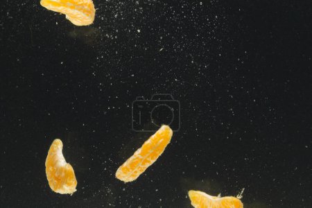 Téléchargez les photos : Gros plan de segments de mandarine tombant dans l'eau avec espace de copie sur fond noir. Fruits, nourriture végétalienne et concept de couleur. - en image libre de droit
