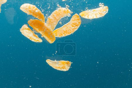 Téléchargez les photos : Gros plan de segments de mandarine tombant dans l'eau avec espace de copie sur fond bleu. Fruits, nourriture végétalienne et concept de couleur. - en image libre de droit