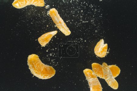 Téléchargez les photos : Gros plan de segments de mandarine tombant dans l'eau avec espace de copie sur fond noir. Fruits, nourriture végétalienne et concept de couleur. - en image libre de droit