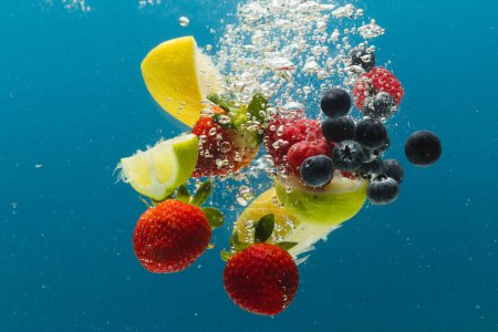 Photo for Close up of berries, lime and lemon slices falling into water with copy space on blue background. Fruit, vegan food and colour concept. - Royalty Free Image