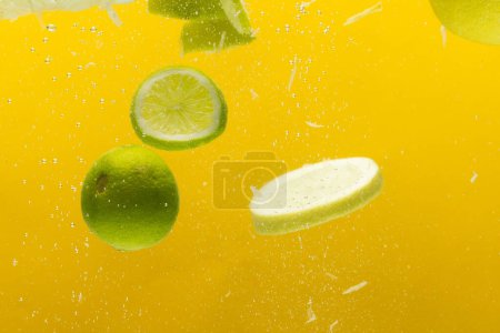Téléchargez les photos : Gros plan de tranches de citron et de citron vert tombant dans l'eau avec espace de copie sur fond jaune. Fruits, nourriture végétalienne et concept de couleur. - en image libre de droit