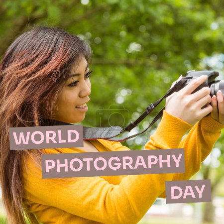 Foto de Día mundial de la fotografía texto en marrón sobre mujer asiática feliz utilizando la cámara en el parque. Celebración global de la campaña fotográfica, imagen generada digitalmente. - Imagen libre de derechos
