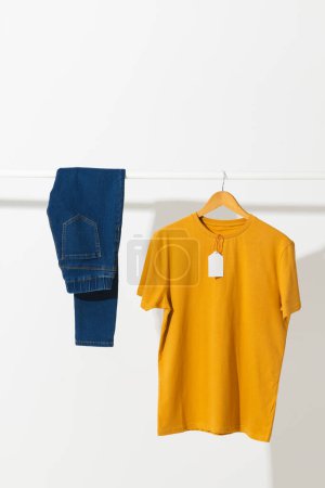 Téléchargez les photos : T-shirt jaune avec étiquette et pantalon en denim sur rail de vêtements avec espace de copie sur fond blanc. Mode, vêtements, concept de couleur et de tissu. - en image libre de droit