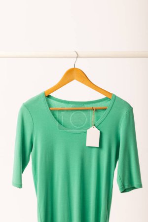 Téléchargez les photos : T-shirt vert avec étiquette sur le cintre suspendu à la rampe de vêtements avec espace de copie sur fond blanc. Mode, vêtements, concept de couleur et de tissu. - en image libre de droit