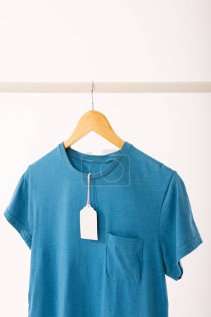 Téléchargez les photos : T-shirt bleu avec étiquette sur le cintre suspendu à la rampe de vêtements avec espace de copie sur fond blanc. Mode, vêtements, concept de couleur et de tissu. - en image libre de droit