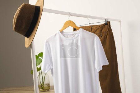Photo for White t shirt, hat and brown trousers hanging from clothes rail with copy space on white background. Fashion, clothes, colour and fabric concept. - Royalty Free Image