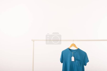 Foto de Camiseta azul con etiqueta en la percha colgando de carril de ropa con espacio de copia sobre fondo blanco. Moda, ropa, color y concepto de tela. - Imagen libre de derechos