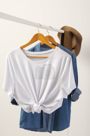 Téléchargez les photos : T-shirt blanc, chapeau et chemise en denim suspendus au rail de vêtements avec espace de copie sur fond blanc. Mode, vêtements, concept de couleur et de tissu. - en image libre de droit