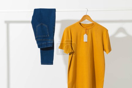 Photo for Yellow t shirt with tag and denim trousers on clothes rail with copy space on white background. Fashion, clothes, colour and fabric concept. - Royalty Free Image