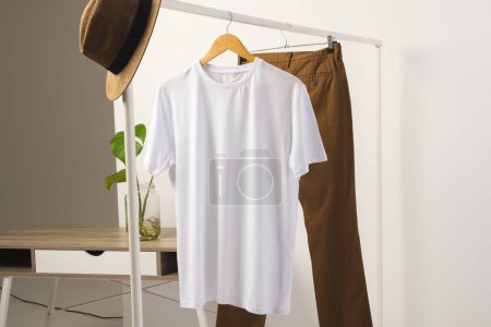 Téléchargez les photos : T-shirt blanc, chapeau et pantalon marron suspendus à la rampe de vêtements avec espace de copie sur fond blanc. Mode, vêtements, concept de couleur et de tissu. - en image libre de droit