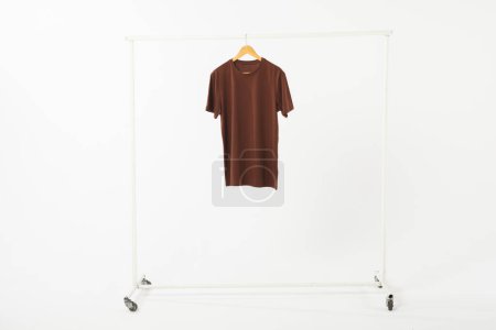 Téléchargez les photos : T-shirt marron sur cintre suspendu au rail de vêtements avec espace de copie sur fond blanc. Mode, vêtements, concept de couleur et de tissu. - en image libre de droit