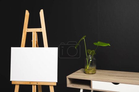 Foto de Lienzo blanco sobre caballete de madera y espacio de copia con escritorio y planta sobre fondo negro. Señalización, espacio de escritura y concepto publicitario. - Imagen libre de derechos