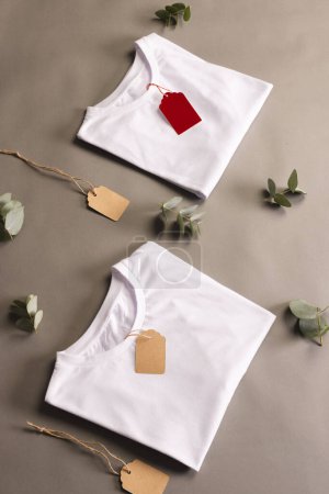 Téléchargez les photos : Chemises pliées blanches avec étiquettes et feuilles avec espace de copie sur fond brun. Mode, vêtements, concept de couleur et de tissu. - en image libre de droit