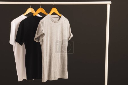 Foto de Tres camisetas en perchas colgando de riel de ropa y espacio de copia sobre fondo negro. Moda, ropa, color y concepto de tela. - Imagen libre de derechos