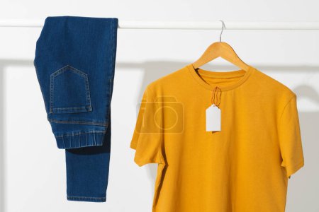 Photo for Yellow t shirt with tag and denim trousers on clothes rail with copy space on white background. Fashion, clothes, colour and fabric concept. - Royalty Free Image