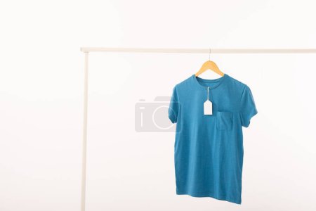 Foto de Camiseta azul con etiqueta en la percha colgando de carril de ropa con espacio de copia sobre fondo blanco. Moda, ropa, color y concepto de tela. - Imagen libre de derechos