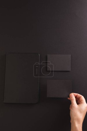 Téléchargez les photos : Image verticale d'un cahier noir et d'une femme caucasienne tenant une carte de papier noir sur fond noir. Concept papier, écriture, texture et matériaux. - en image libre de droit