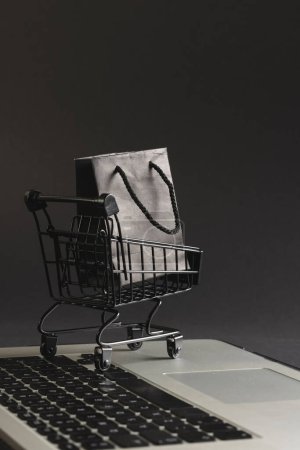 Photo for Vertical image of laptop and trolley with bag and copy space on black background. Cyber monday, cyber shopping, retail, technology, electronic device and communication concept. - Royalty Free Image