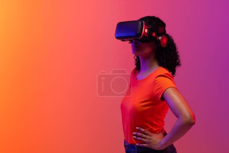 Foto de Mujer Biracial usando auriculares vr sobre fondo de neón de color rosa a naranja. Tecnología, redes digitales, conexiones globales y metáfora de la comunicación. - Imagen libre de derechos