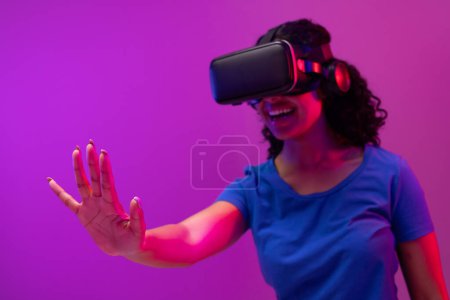 Téléchargez les photos : Femme Biracial utilisant vr casque et pointant sur fond rose fluo à violet. Technologie, réseaux numériques, connexions mondiales et métaverse de la communication. - en image libre de droit