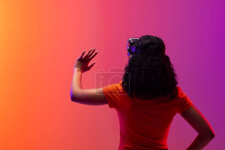 Foto de Mujer biracial usando auriculares vr y apuntando sobre fondo de color rosa neón a naranja. Tecnología, redes digitales, conexiones globales y metáfora de la comunicación. - Imagen libre de derechos
