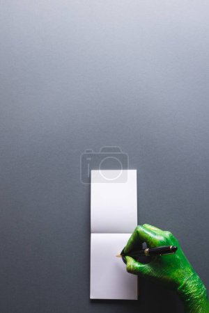 Téléchargez les photos : Image verticale de mains de monstre vert écriture carnet avec espace de copie sur fond gris. Halloween, concept de tradition et célébration. - en image libre de droit
