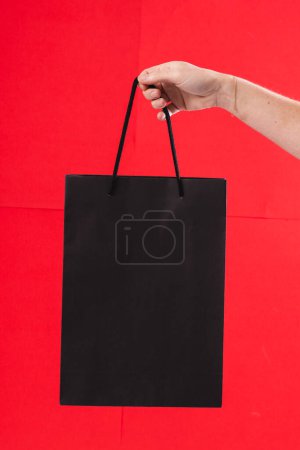 Foto de Mano de mujer caucásica sosteniendo bolsa de regalo negro con espacio de copia sobre fondo rojo. Viernes negro, lunes cibernético, compras, compras cibernéticas, ventas, venta al por menor y el concepto de envío. - Imagen libre de derechos