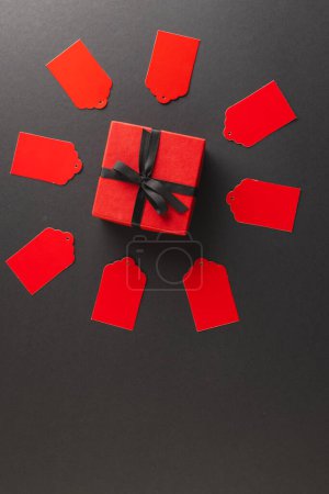 Photo for Vertical image of red gift tags in circle and red gift box with copy space over black background. Cyber monday, black friday, online shopping, shipping and global connections concept. - Royalty Free Image